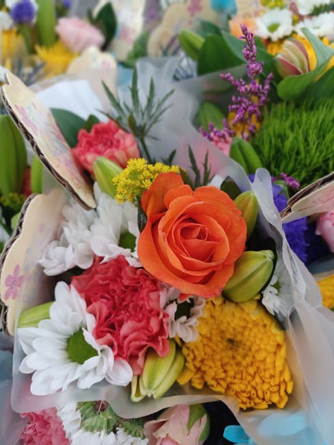 A vibrant bouquet featuring a central orange rose surrounded by white daisies, pink carnations, yellow chrysanthemums, and purple lavender sprigs, all arranged with lush green foliage and wrapped in d