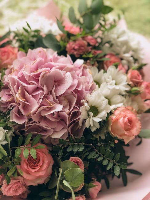 A detailed close-up of a floral bouquet featuring soft pink hydrangeas with large, ruffled petals, white daisies with yellow centers, and pale pink roses with tightly layered petals. The arrangement i