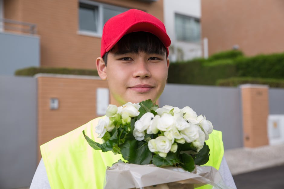 A young male delivery person wearing a red cap and a high-visibility yellow vest holds a bouquet of fresh white flowers wrapped in white paper. The bouquet features delicate petals and green foliage, 