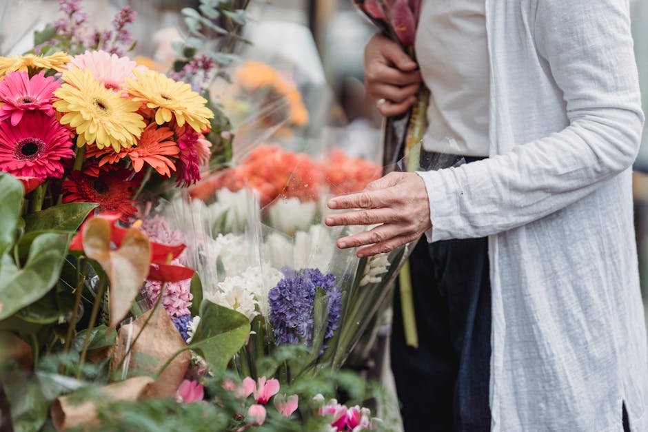 A person selecting fresh flowers at a flower stall, with an assortment of vibrant blooms including pink, yellow, purple, and white gerbera daisies and other flowers, wrapped in clear plastic and arran