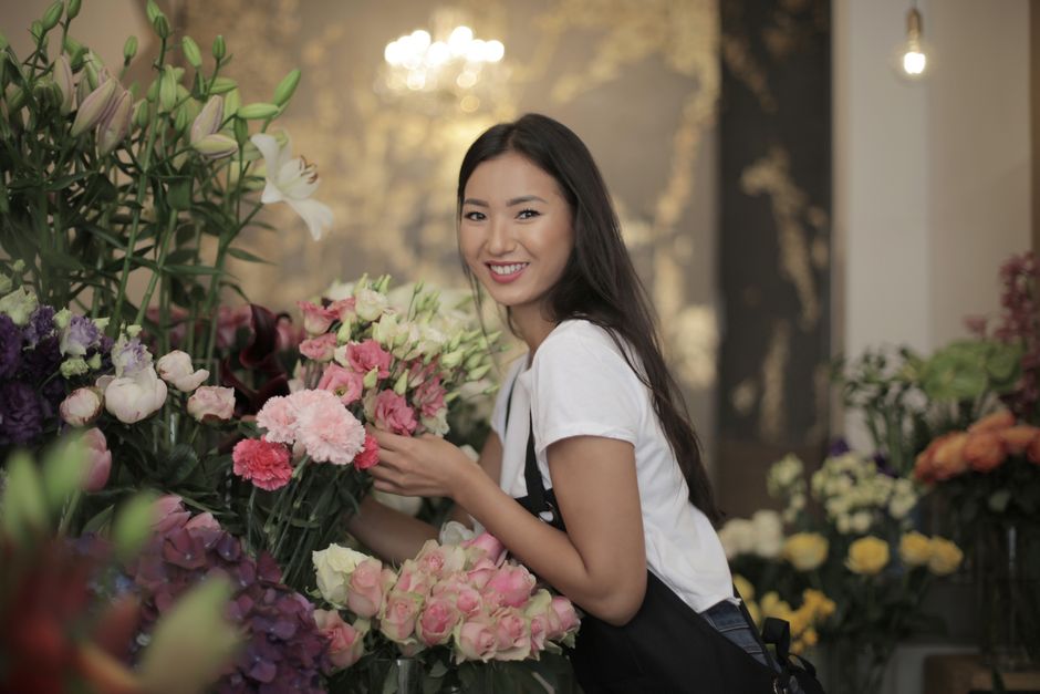 A young woman with long dark hair and a white t-shirt smiling while holding a bouquet of pink and white flowers in a flower shop. She is surrounded by an assortment of fresh flowers in various colors,
