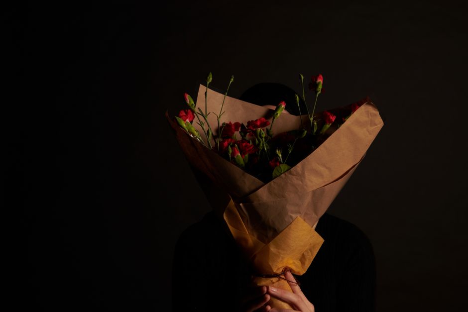 A person holding a bouquet of fresh red flowers, including various blossoms with green stems and leaves, wrapped in brown paper and tied with a ribbon. The bouquet is positioned in front of their face