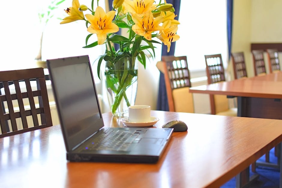 A clear glass vase filled with vibrant yellow lilies with dark stamen details, arranged on a polished wooden conference table. The bouquet features fresh, open blooms and green stems, wrapped in minim