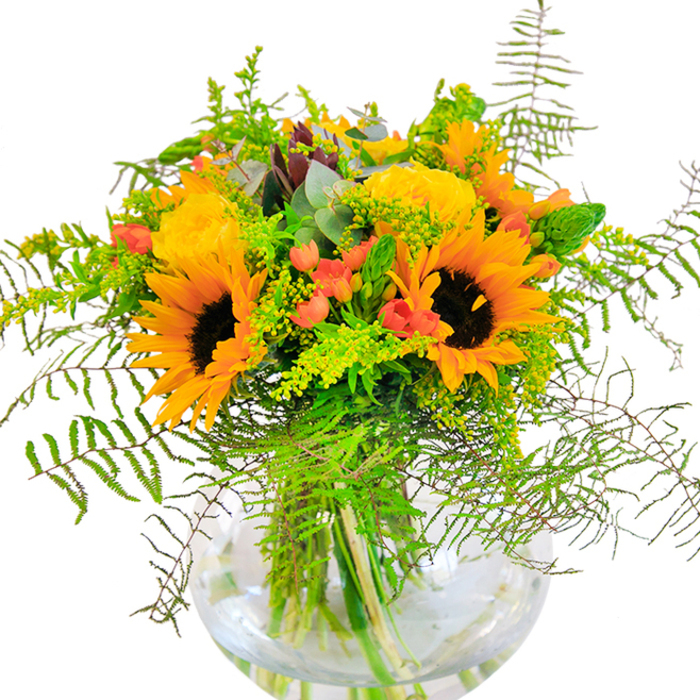 The vibrant spirit of Bracknell is beautifully encapsulated in this abundant floral arrangement, a harmonious blend of color and texture crafted by skilled local hands. At its core, bold sunflowers radiate with their iconic yellow-orange petals and contrasting coffee-brown centers, evoking the golden sunlight that bathes the borough's parks. Nestled among them, sunny yellow garden roses unfurl in elegant spirals, their lush blooms adding softness and classic sophistication. The sun-warmed hues are accentuated by glossy red Hypericum berries, whose playful bursts of color glisten like berries glimpsed at harvest along Ascot Road. Fern fronds cascade gently, lending a touch of wild movement and elevating the sense of natural abundance, while filmy sprays of solidago add piquant flecks of gold reminiscent of Bracknell's late summer hedgerows. The presence of eucalyptus, with its distinctive blue-green leaves, infuses the bouquet with a refreshing countryside vibe, nodding to the town's leafy walking routes and gardens. Arranged in a round, clear glass vase, the green stems appear artful and tidy, floating in crystal-clear water that sparkles beneath the well-chosen blooms. This bouquet embodies joy and optimism-ideally placed as a centerpiece for birthdays, anniversaries, or to simply bring a radiant cheer into a local home. As your nearby florist, each element is selected to echo the warmth and lively character of Bracknell, promising an uplifting delivery for any occasion.