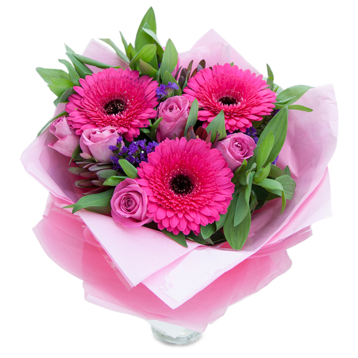 I picture this bouquet as a thoughtful, tactile gift - three vivid fuchsia Gerbera daisies commanding attention with their broad, radiating petals and dark centres that provide a striking contrast, surrounded by roses in a gentle lavender-pink, each bloom still slightly furled in that fresh, promising stage. Between them, sprigs of deep purple statice peek out, their delicate, papery clusters adding playful texture and a subtle contrast that deepens the pink palette. The greenery is generous and purposeful: glossy salal-like leaves create a full, green backdrop while touches of eucalyptus lend a silvery whisper and a faint herbal note. The flowers are cradled in an inner layer of soft, matte pale pink tissue paper and wrapped again in a translucent fuchsia-pink cellophane that shimmers when it catches light; the cellophane's slight crinkle and the neat folding speak of careful handling and a desire to present the flowers like a cherished gift. At the base the top rim of a clear glass vase is visible, hinting at freshness and immediate display readiness. In gentle daylight the petals read as velvety and tactile, the greens crisp and slightly dewy; the scent would be a light mingling of rose perfume and the green sweetness of Gerberas. I imagine this bouquet arriving to a home in Bracknell - perhaps after a visit to Lily Hill Park or a stroll through The Lexicon - to mark a birthday, an anniversary, or simply to bring unexpected joy and comfort.